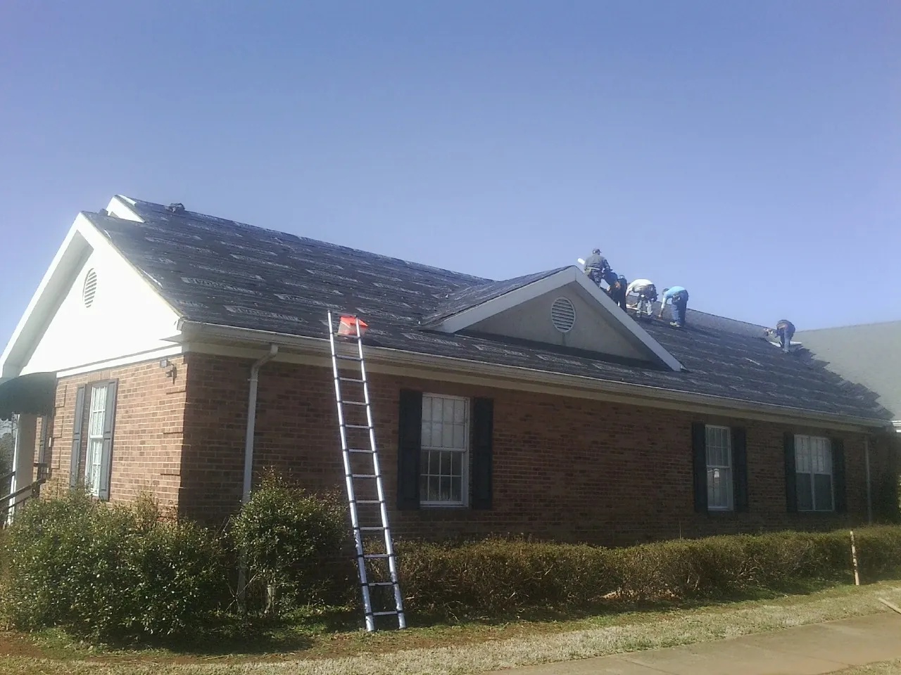 Crew tearing shingles off a brick ranch roof with a ladder set — Emergency Roof Repair work by our crew in Auburn Hills