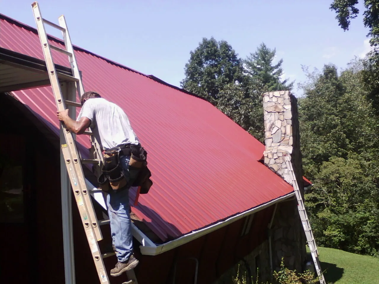 Roofer on a ladder beside a red metal roof and stone chimney — Soffit & Fascia Repair by Roofing Brothers in Auburn Hills